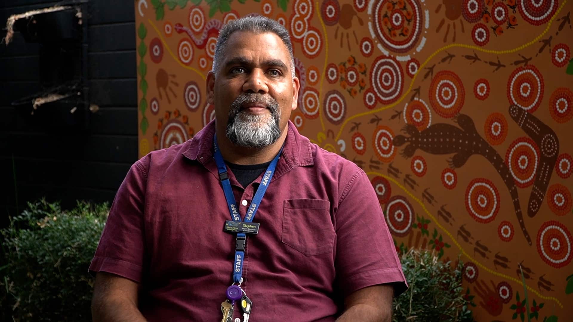 A man - Stephen Morrison - sits in front of a wall featuring a detailed Aboriginal artwork.