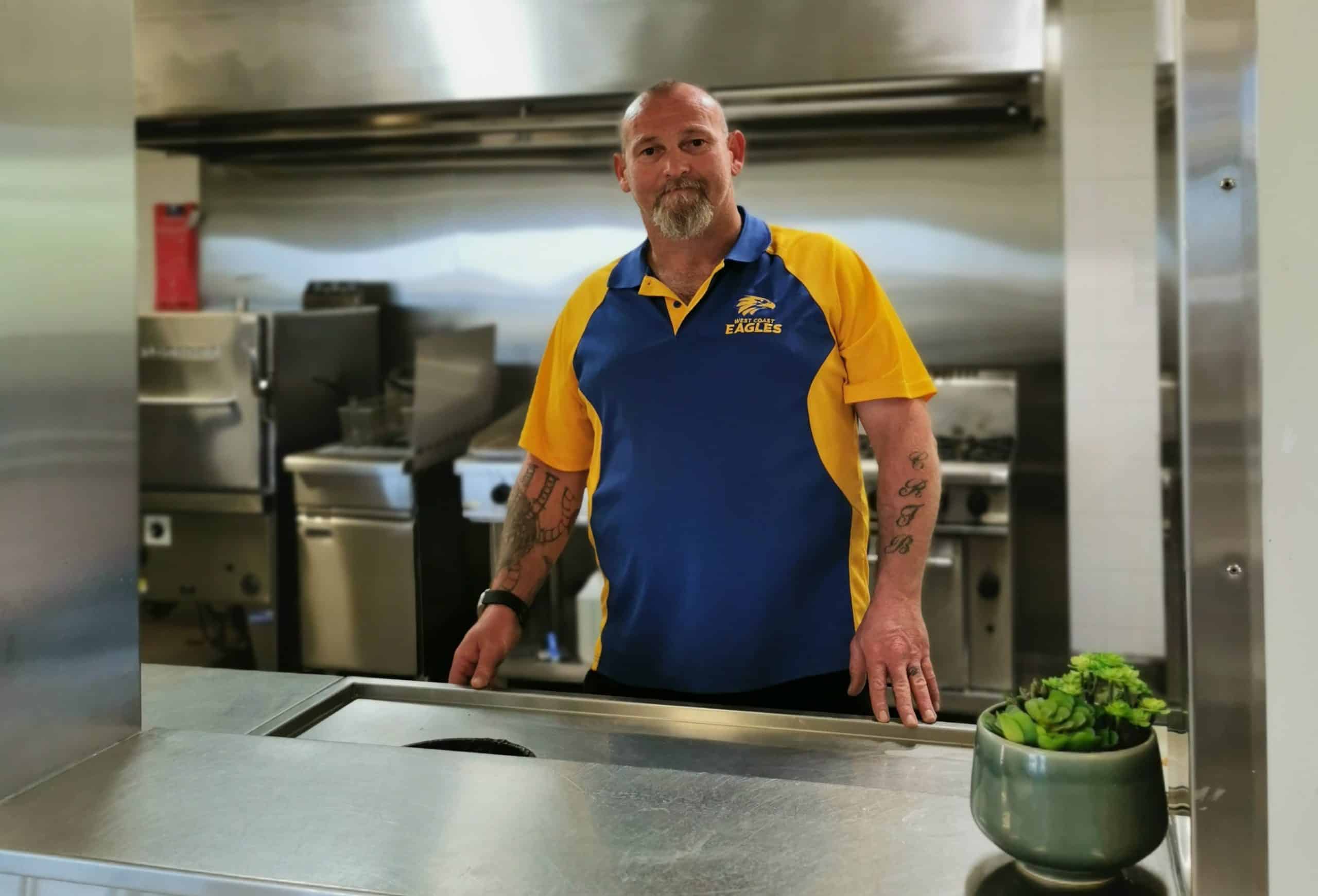A path to recovery: A man in a blue and yellow polo shirt featuring the West Coast Eagles team emblem stands in an industrial kitchen.