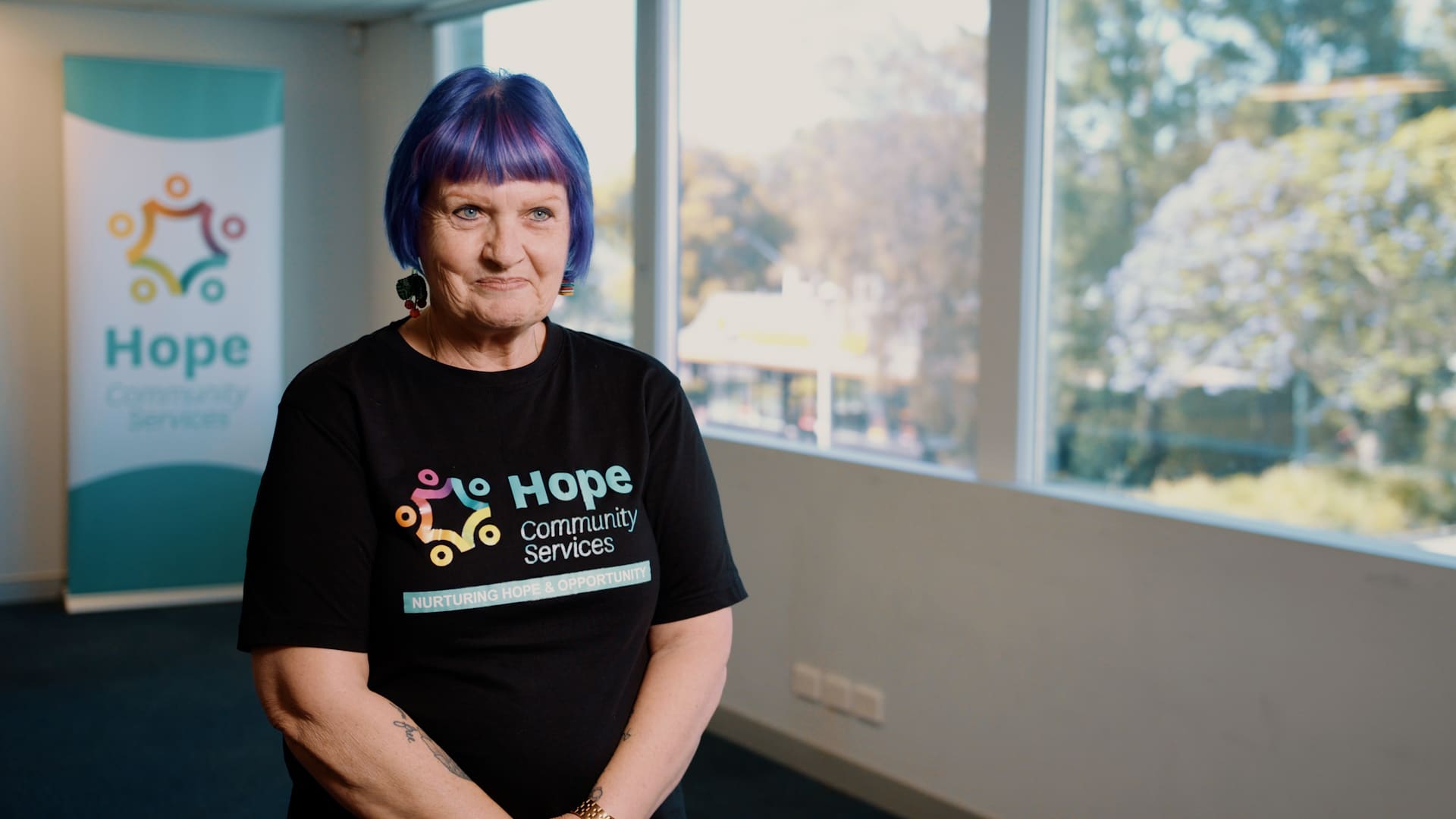 Profile picture of Leeanne Bates - a woman with purple hair who is wearing a black t-shirt with Hope Community Services written on it.
