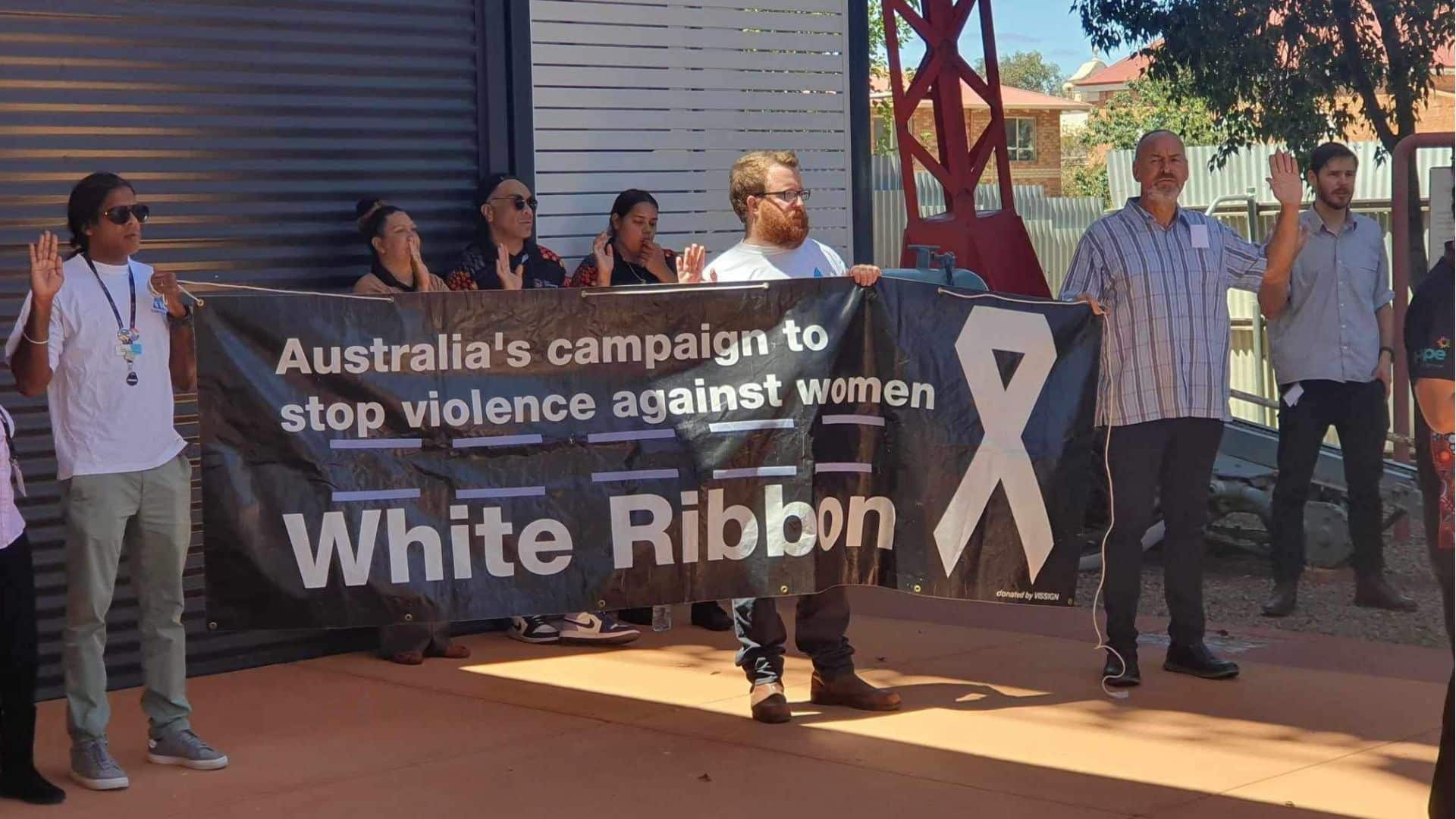 FDV Pledge: A group of men and women stand with their hands raised, palms out, looking solemn. Two are holding a banner that reads: "Australia's campaign to stop violence against women."
